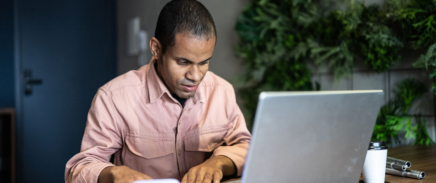 A blind man in a soft pink shirt is seated at a desk in front of an open laptop and cellphone, his folded white cane and a takeaway coffee cup sit alongside