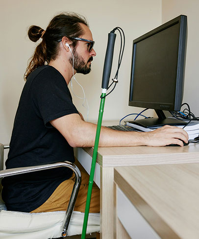 A person with long hair in bun and dark glasses works at a computer screen with his cane resting on the desk next to him.